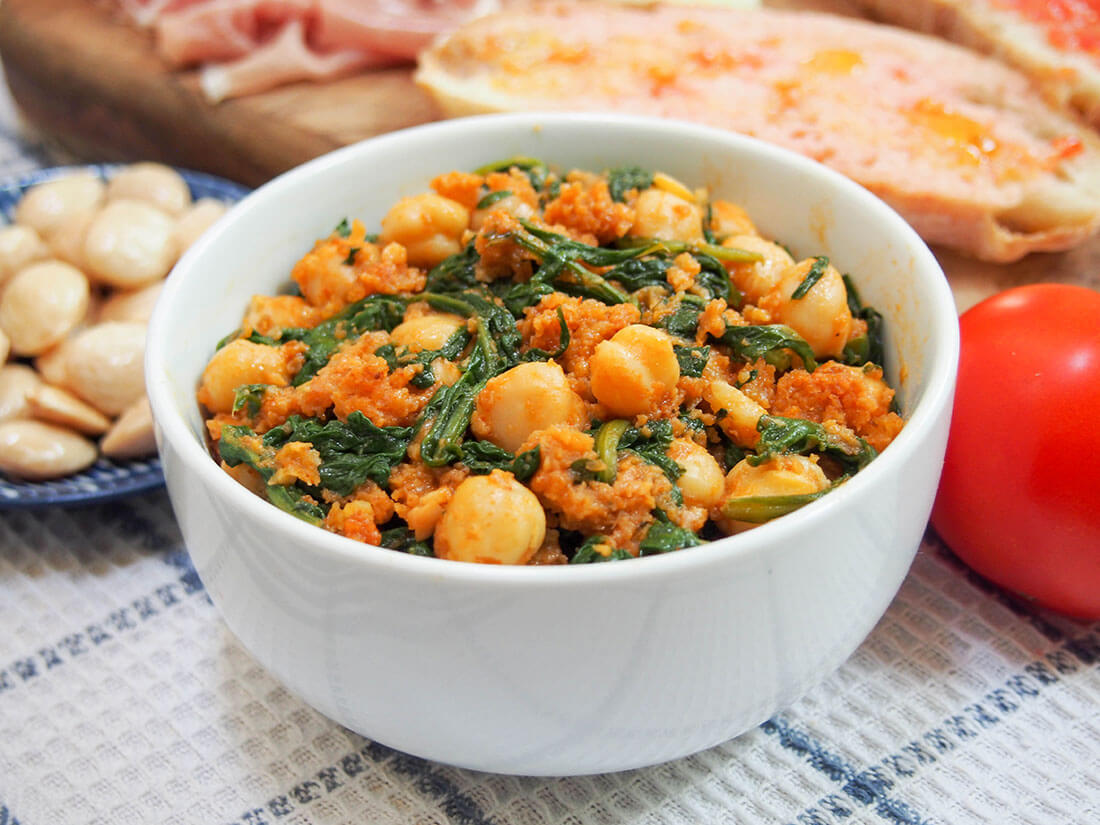 Spanish chickpeas and spinach in bowl with pan con tomate and almonds behind
