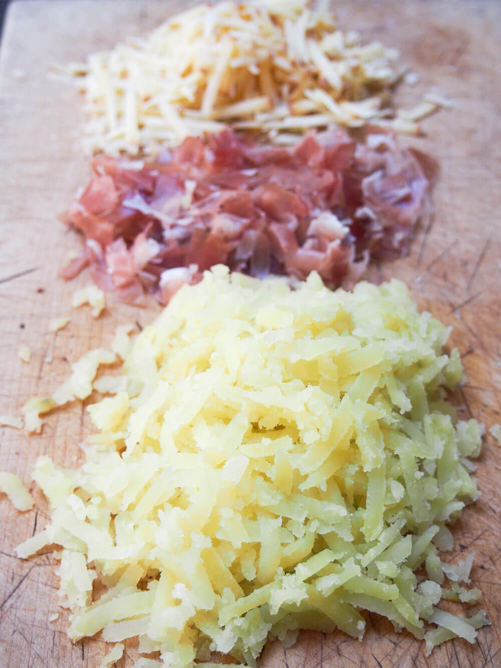 stacks of grated potato, chopped speck and grated cheese on chopping board