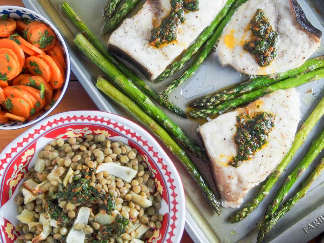 Baked swordfish and asparagus on baking sheet with lentils and chermoula and carrot salad sides in bowls