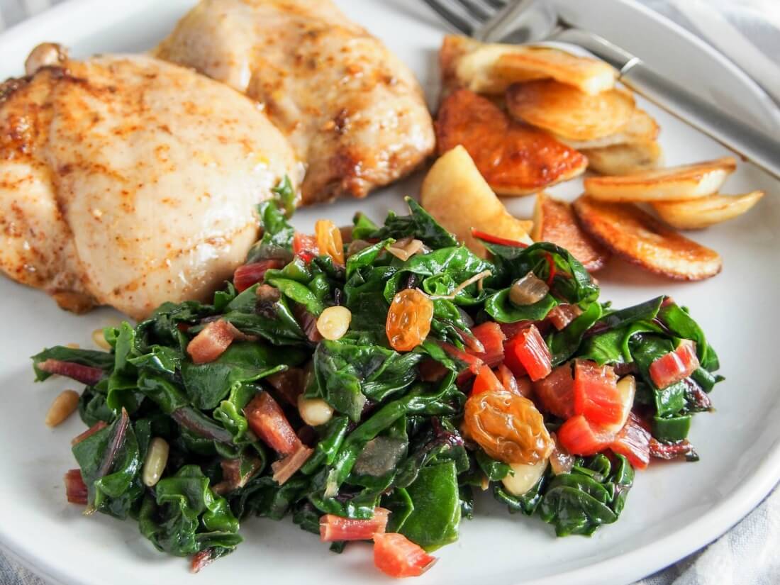Catalan-style sauteed Swiss chard with raisins and pine nuts on plate with chicken and pan-fried potatoes behind