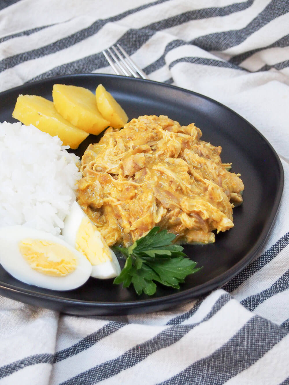 Aji de gallina (Peruaanse kip) met rijst, ei en aardappelen ernaast, alles op een zwart bord