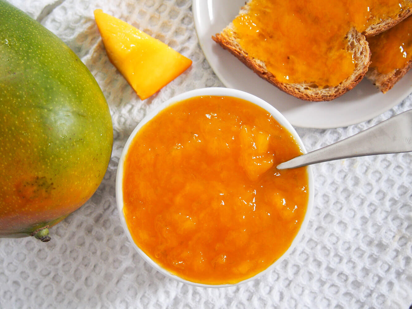 bowl of mango jam with spoon in it with jam on toast to one side and mango to other