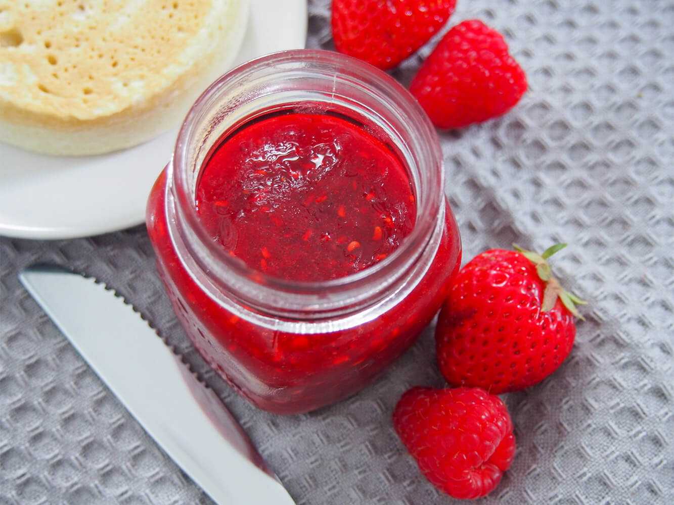 strawberry raspberry jam in jar from overhead with fruit to side and crumpet on plate in corner