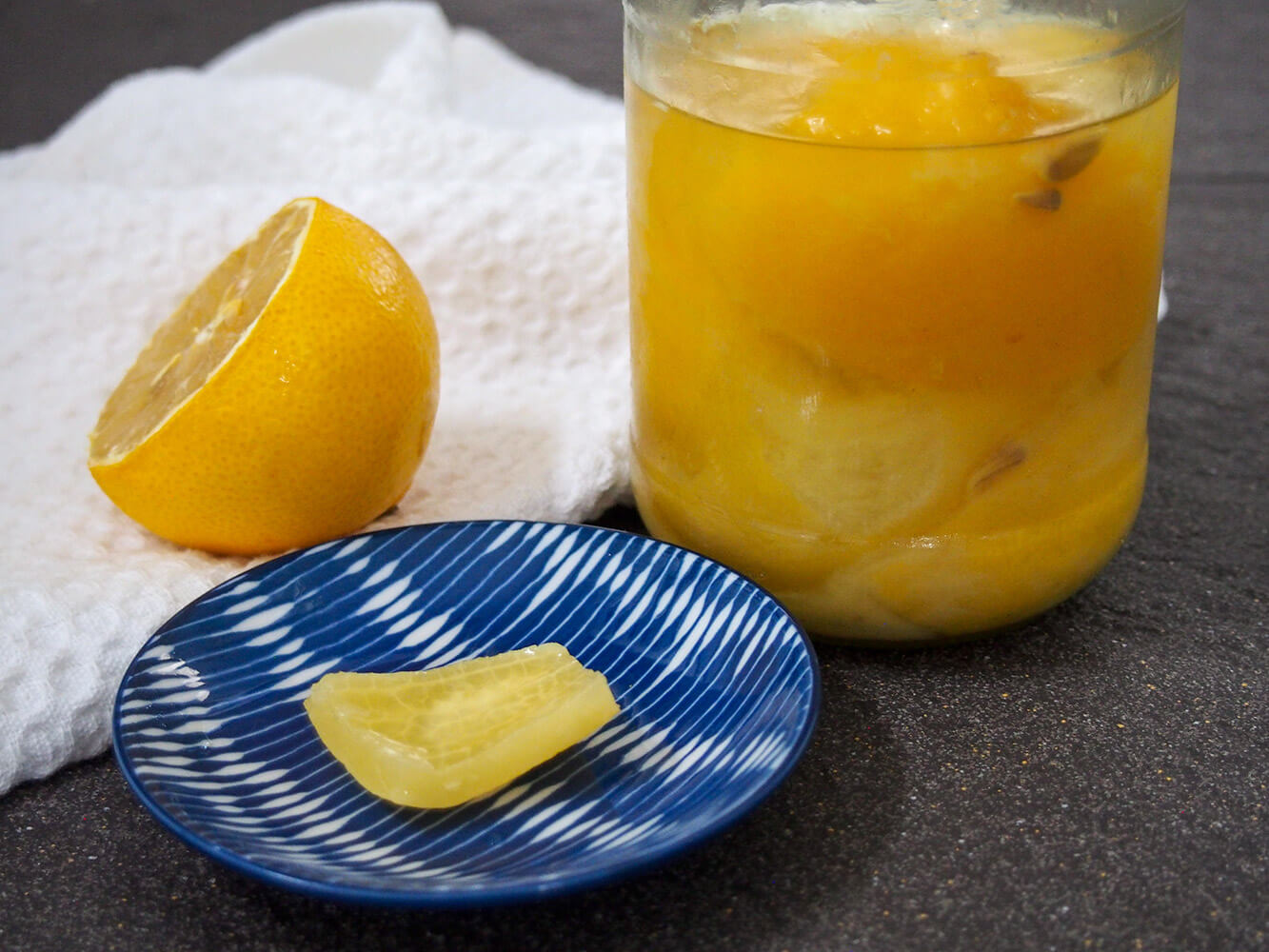 piece of preserved lemon in front of jar with more with fresh lemon half to side