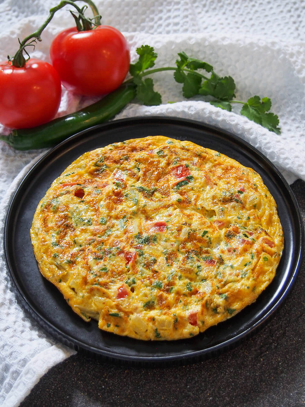 masala omelette on plate with tomato, chili and cilantro behind