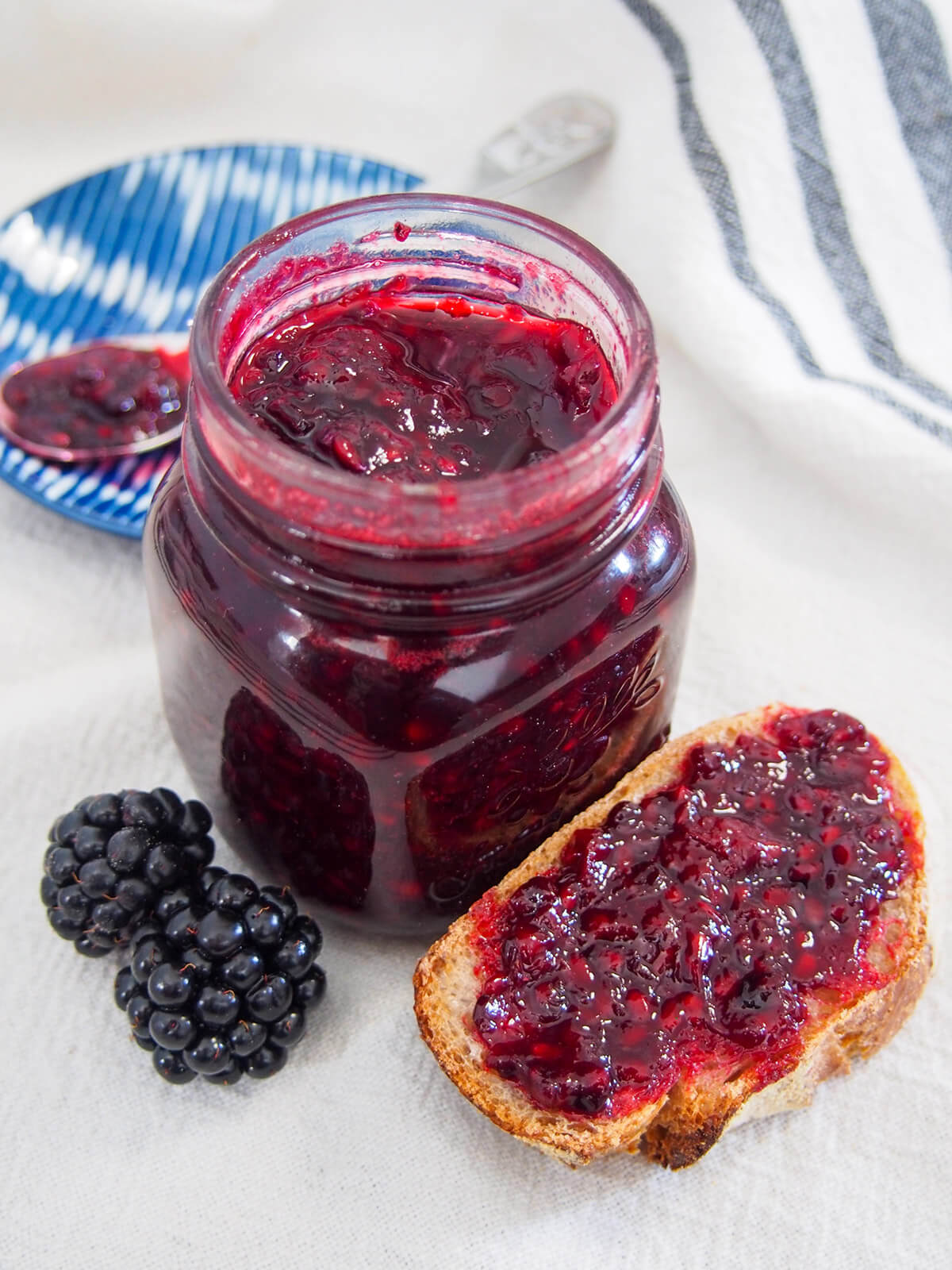 jar of blackberry jam with some on bread to one side and blackberries in front on other side