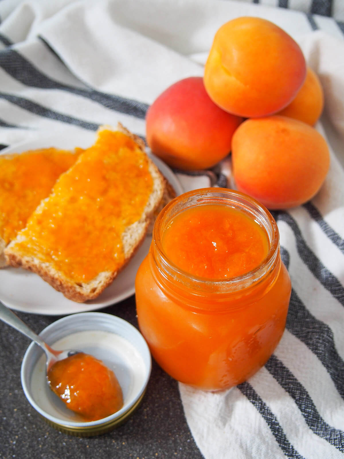 jar or apricot jam with spoon filled with jam to side, toast and stack of apricots behind