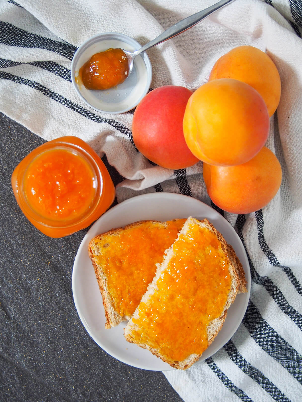 toast spread with apricot jam on plate with jar of jam to side and stack of apricots above