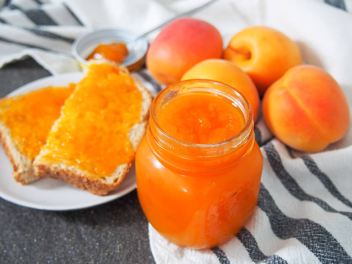 jar of apricot jam with bread and jam and fresh apricots behind jar