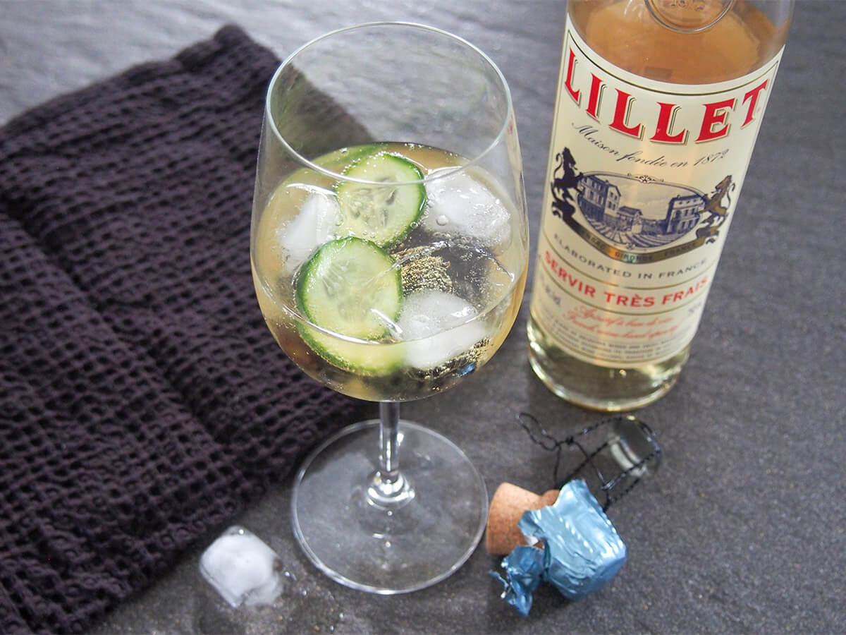 glass of lillet spritz with cucumber in glass and bottle of lillet to side and black cloth on other side