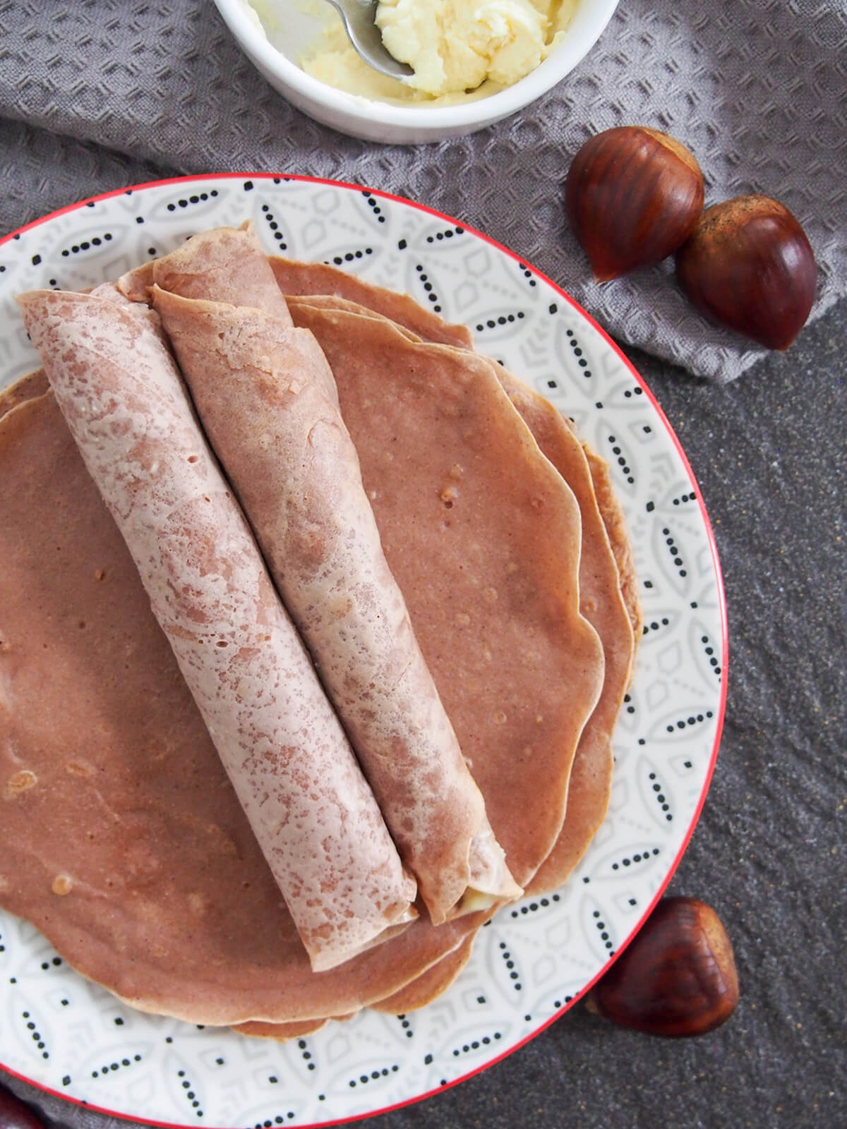 two rolled up chestnut crepes on top of others laid out flat on plate viewed from overhead with some mascarpone in bowl above