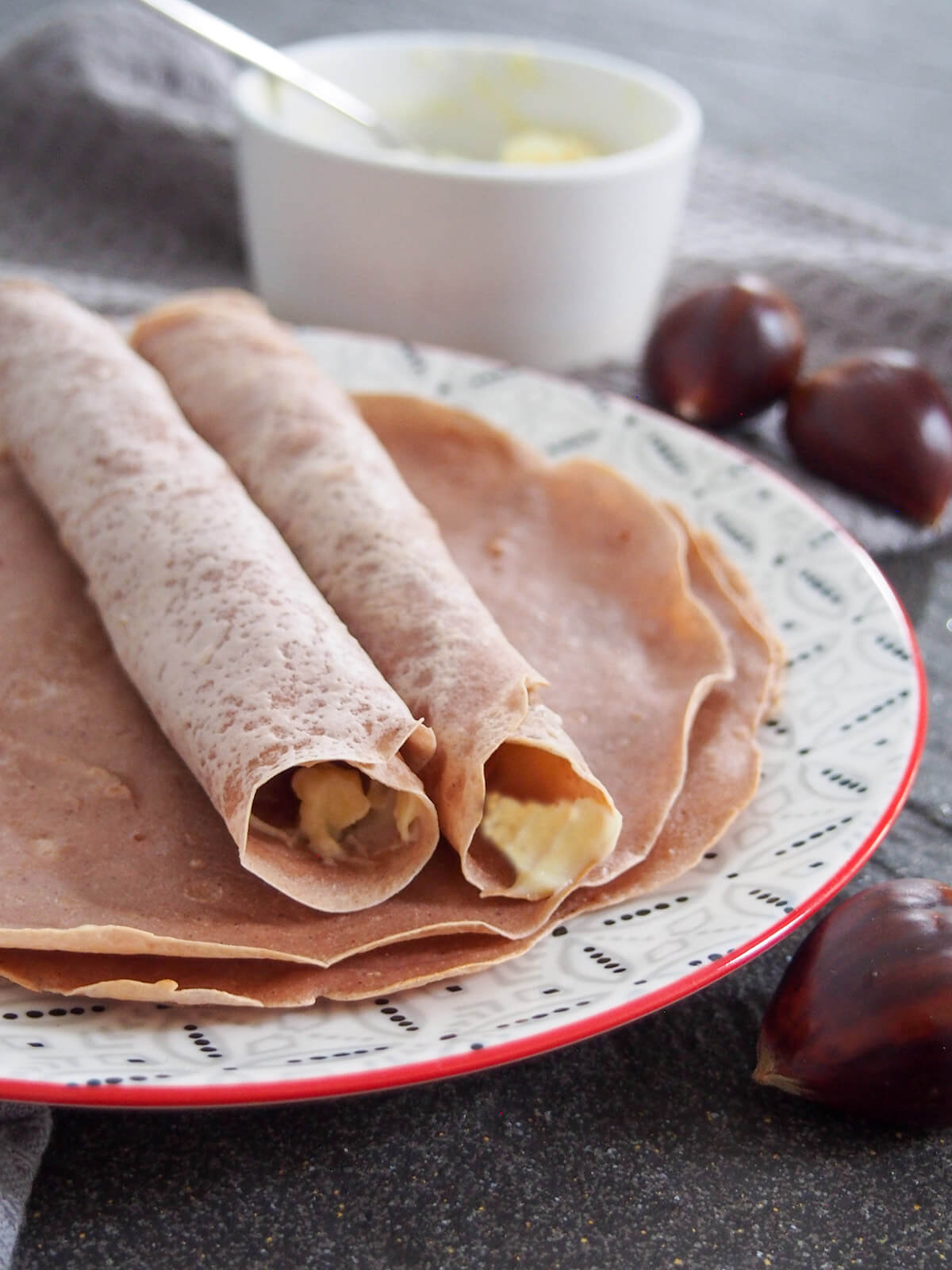 two chestnut crepes rolled up and part view of others under them on plate, small bowl with spoon in it behind