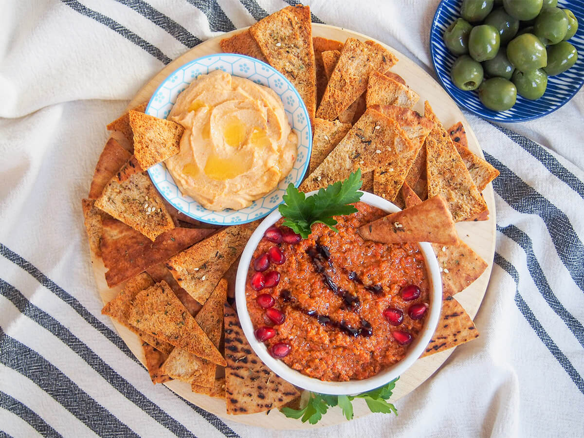 board with baked pita chips, two bowls of dips and olive to side