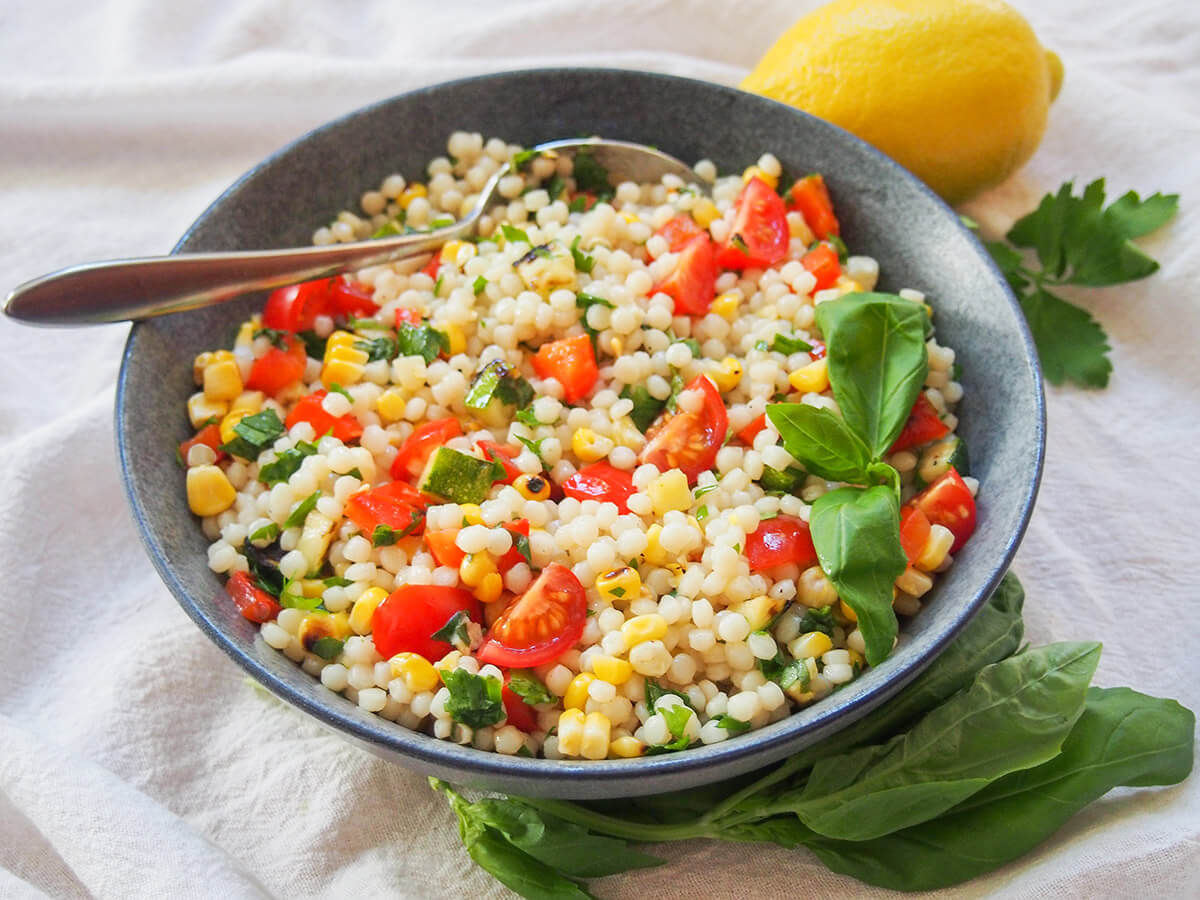 bowl of Israeli couscous salad with grilled vegetables with herbs below bowl and spoon in back of bowl