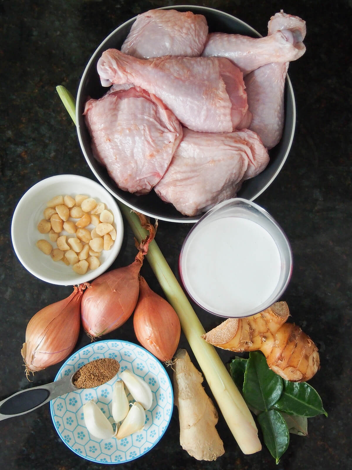 ingredients for opor ayam bowl of chicken pieces, with lemongrass stalk, shallots, galangal, lime leaves, garlic and coconut milk around.
