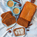 overhead view of loaf of ontbijtkoek Dutch spice cake with slices on plate and small plate with a slice with butter, vups of coffee between.