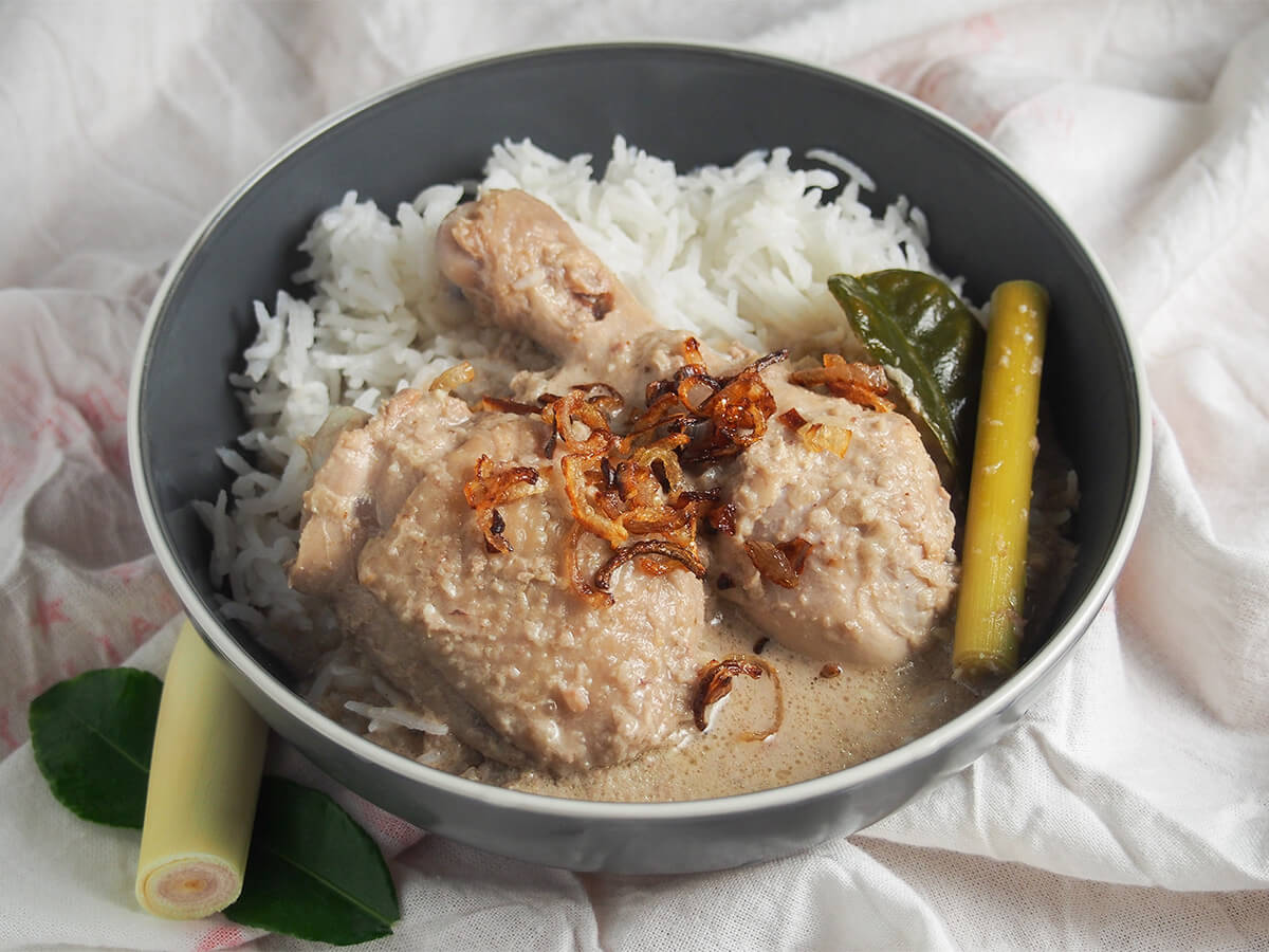 bowl of opor ayam Indonesian coconut chicken with lemongrass stalk and lime leaves to side of bowl.