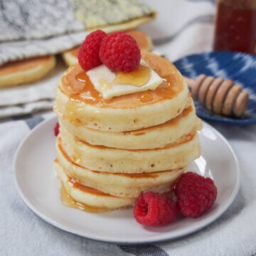 stack of Scotch pancakes in plate with raspberries on top and to side.