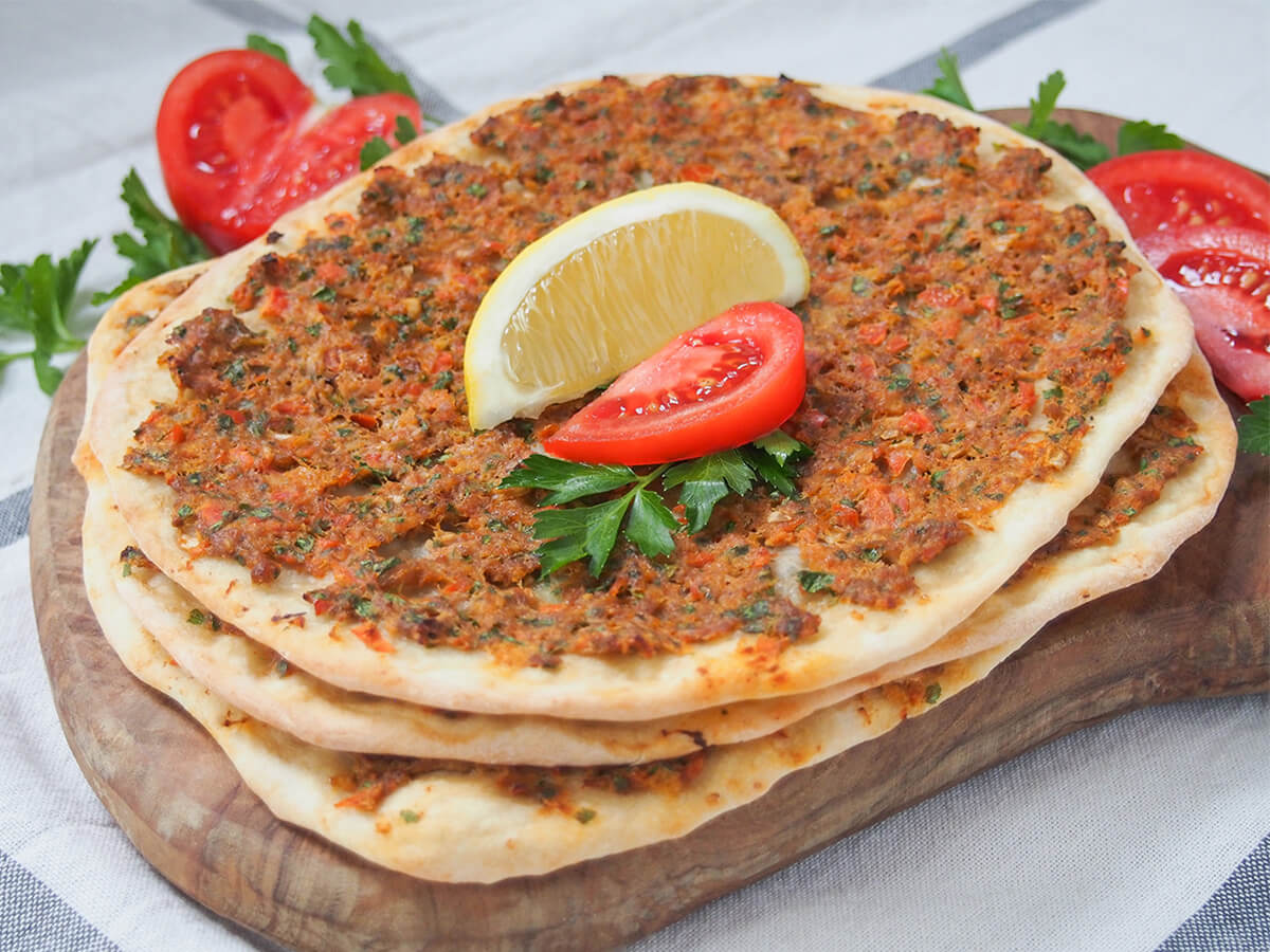 three lahmacun Turkish pizza stacked on top of each other on wooden board with lemon and tomato wedges on top.