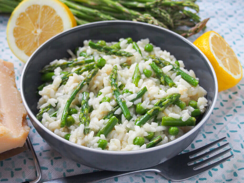 A gray bowl of creamy risotto topped with asparagus and peas. The bowl is placed on a light-colored, patterned tablecloth with a fork beside it. In the background, there are lemon halves, fresh asparagus spears, and a wedge of cheese.