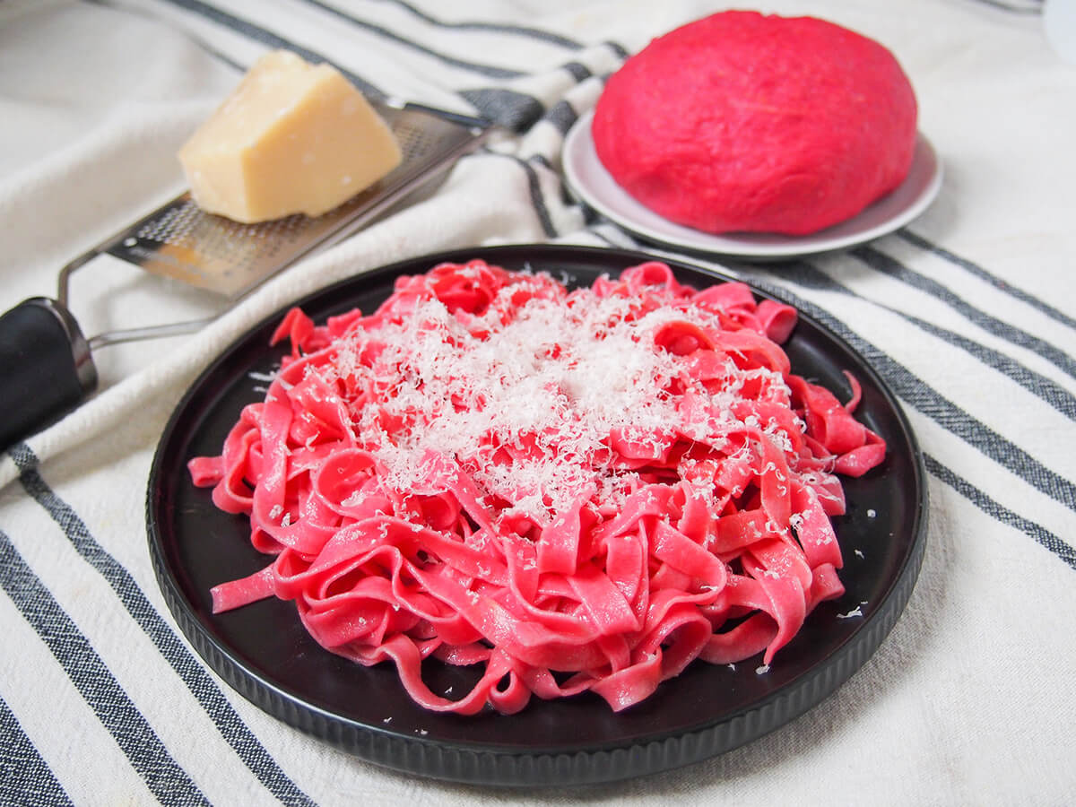 plate of beet pasta plate of beet pasta topped with parmesan with block of cheese and beet pasta dough behind.