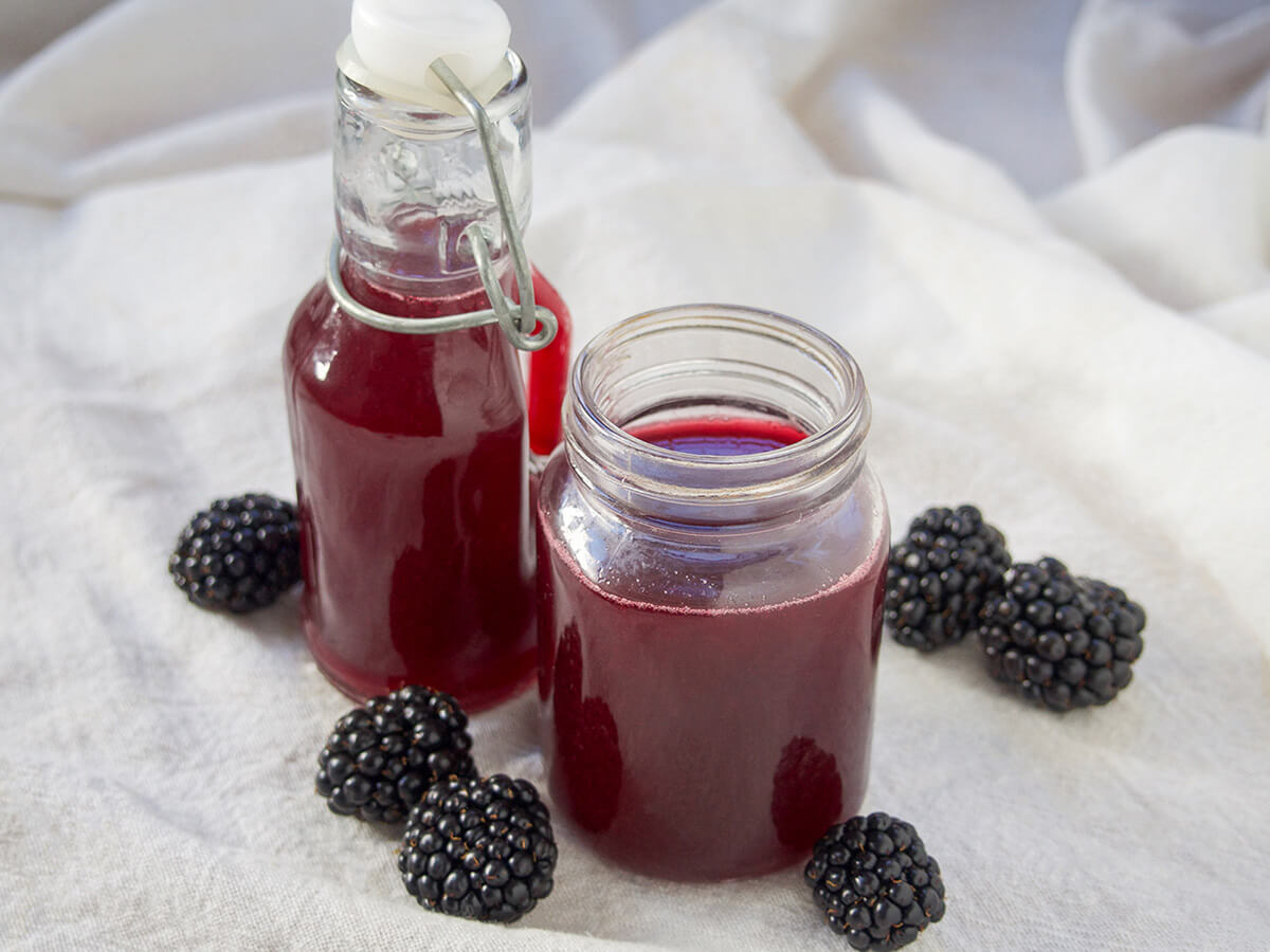 bottle and jar of blackberry simple syrup. jar and small bottle of blackberry simple syrup with blackberries either side.