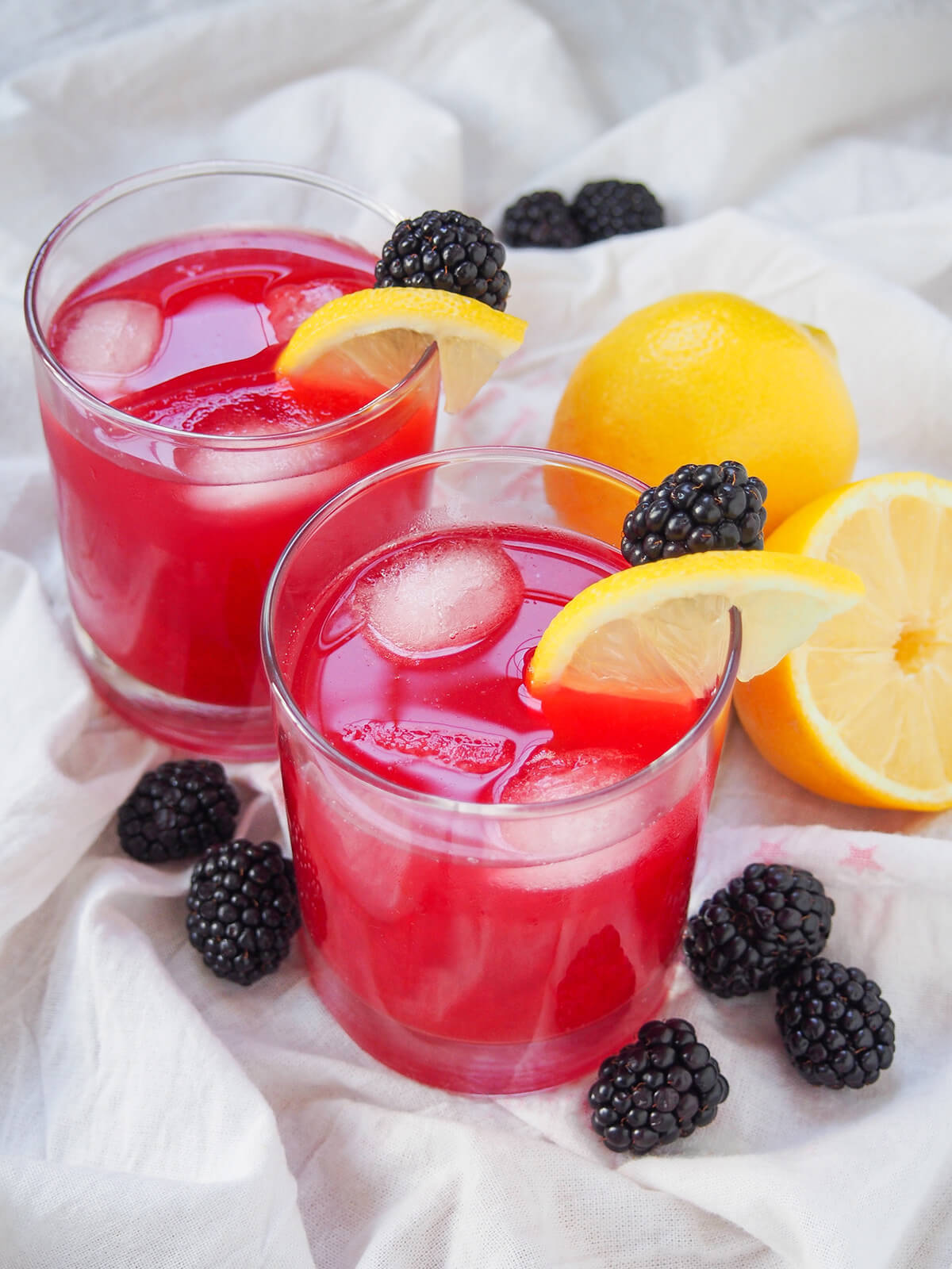 two glasses of blackberry lemonade with blackberries to side of glass and lemon behind.