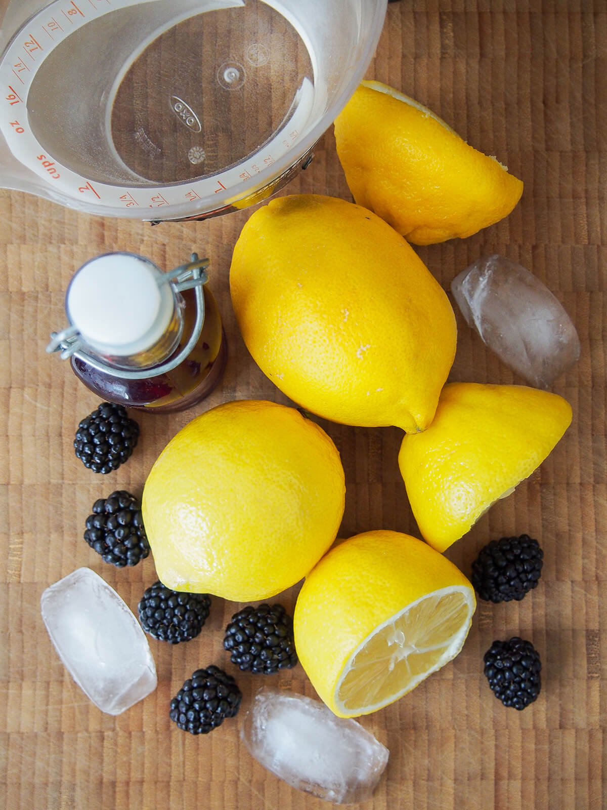whole and cut lemons, blackberries, ice and bottle of syrup on chopping board.