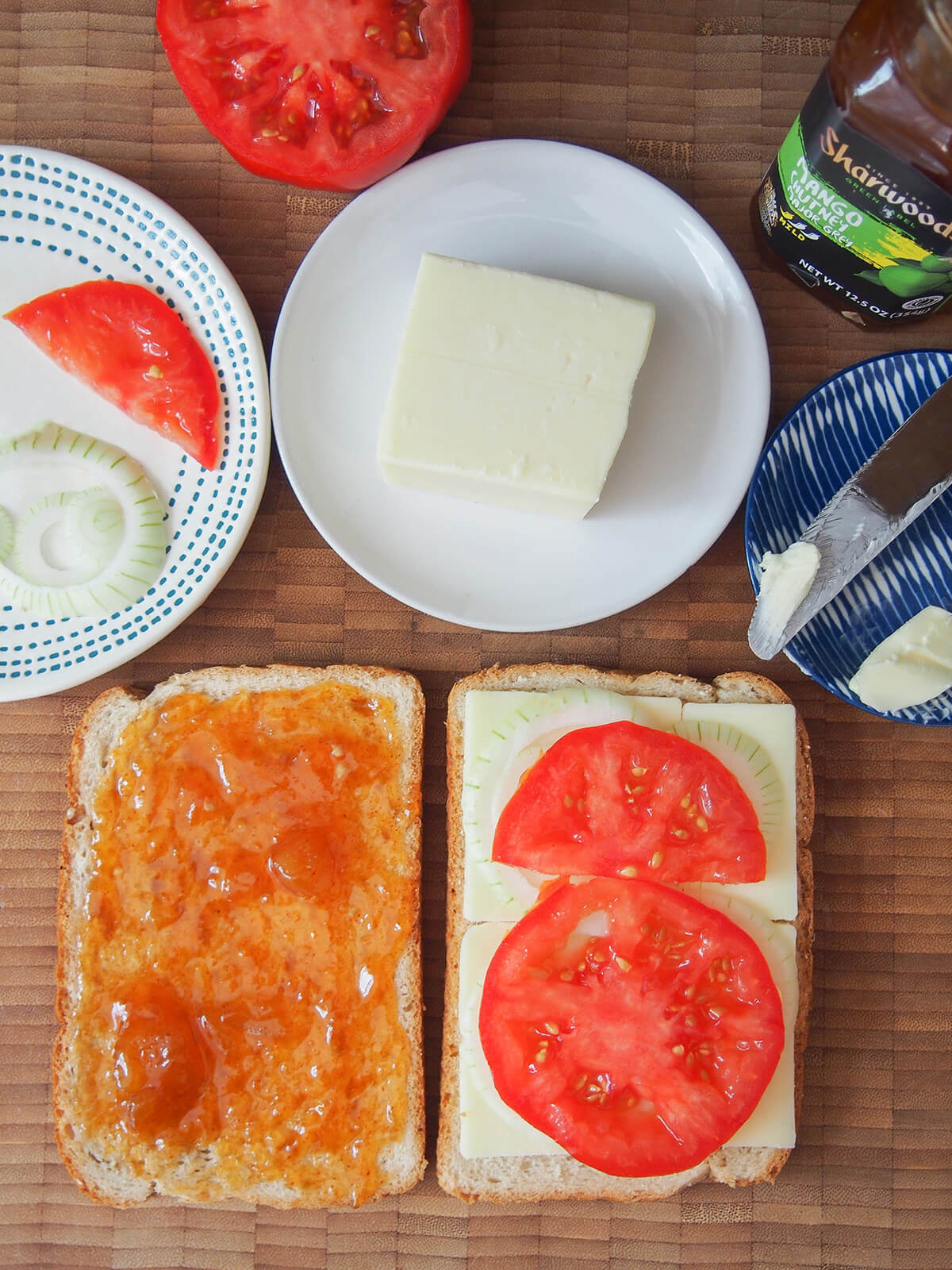 making South African grilled cheese sandwich, braaibroodjie one piece of bread topped with chutney and another topped with cheese, tomato and onion with plates with ingredients aboce.