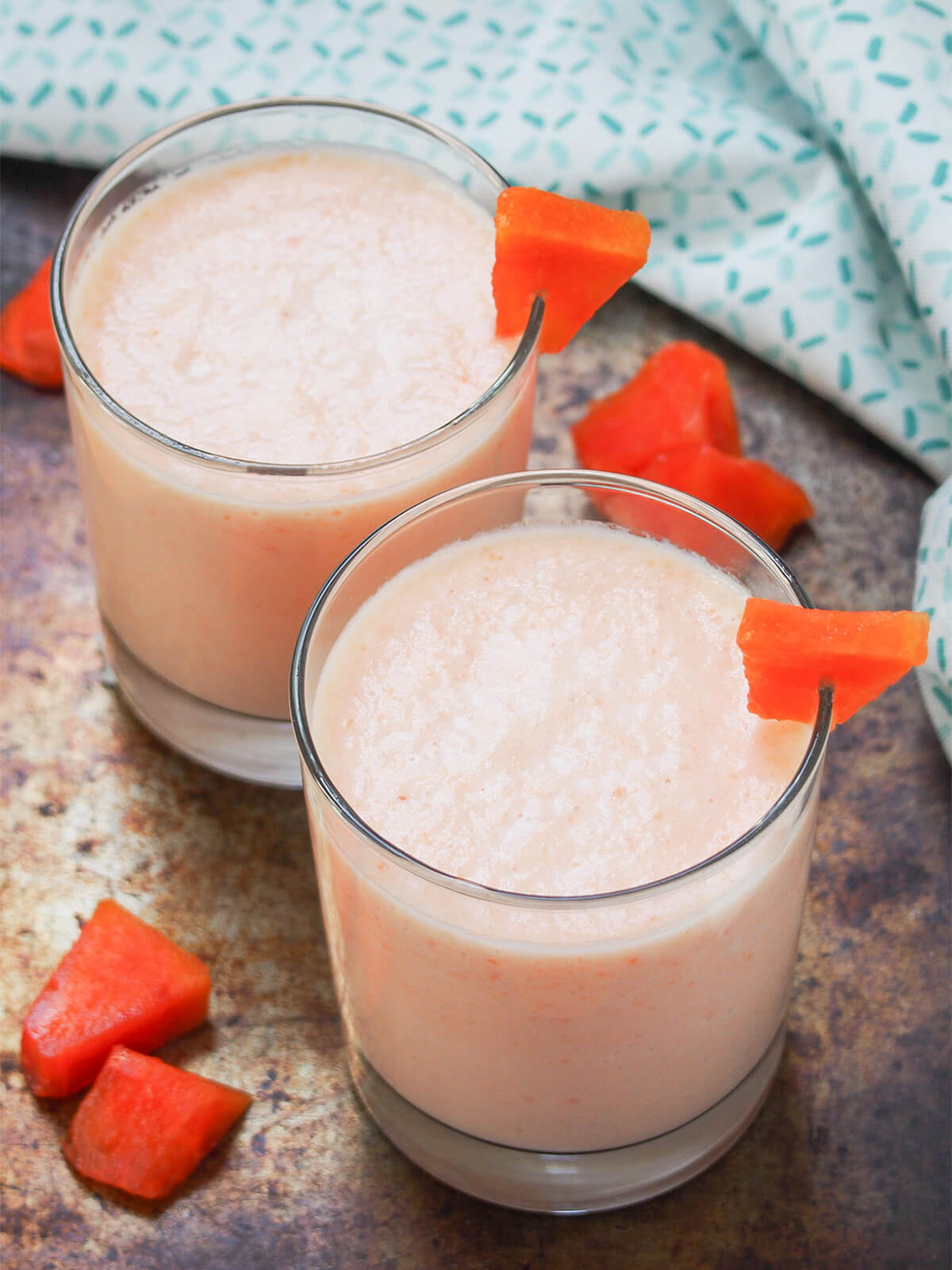 This papaya milkshake is creamy, fruity, cool and delicious. It's easy to make, refreshing and the delicate tropical flavor is perfect for a warm day (or to imagine you're somewhere warm). two glasses of papaya milkshake garnished with pieces of papaya and also papaya to side.