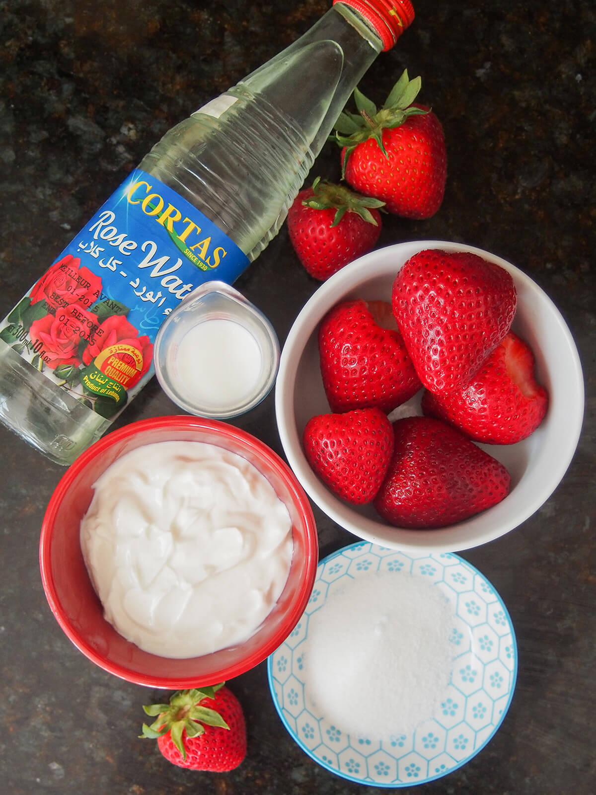 bottle of rosewater, bowls of strawberries and yogurt with some milk and sugar to side.