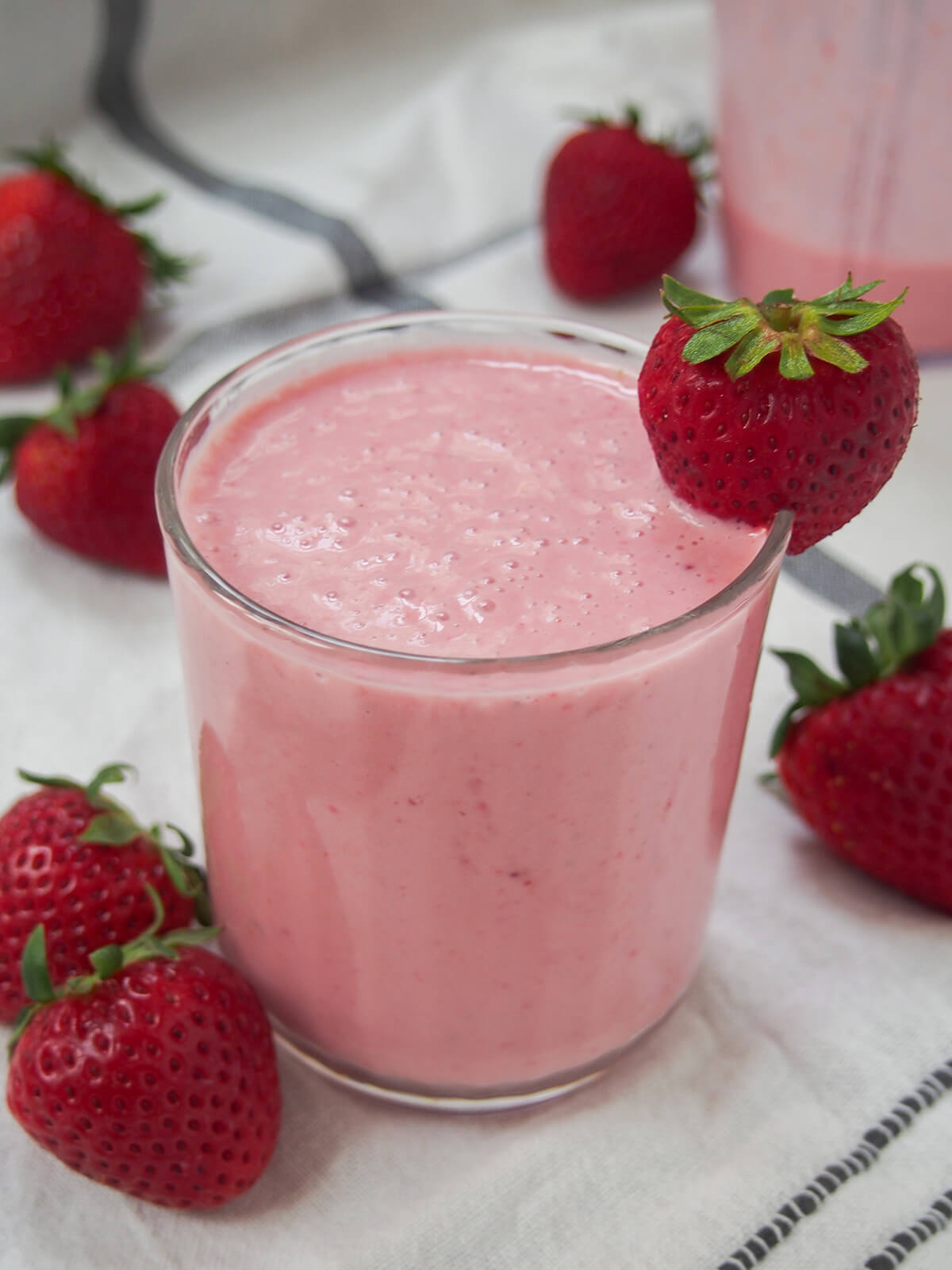 side view of glass of strawberry lassi with strawberries and blender jug with small amount left behind.