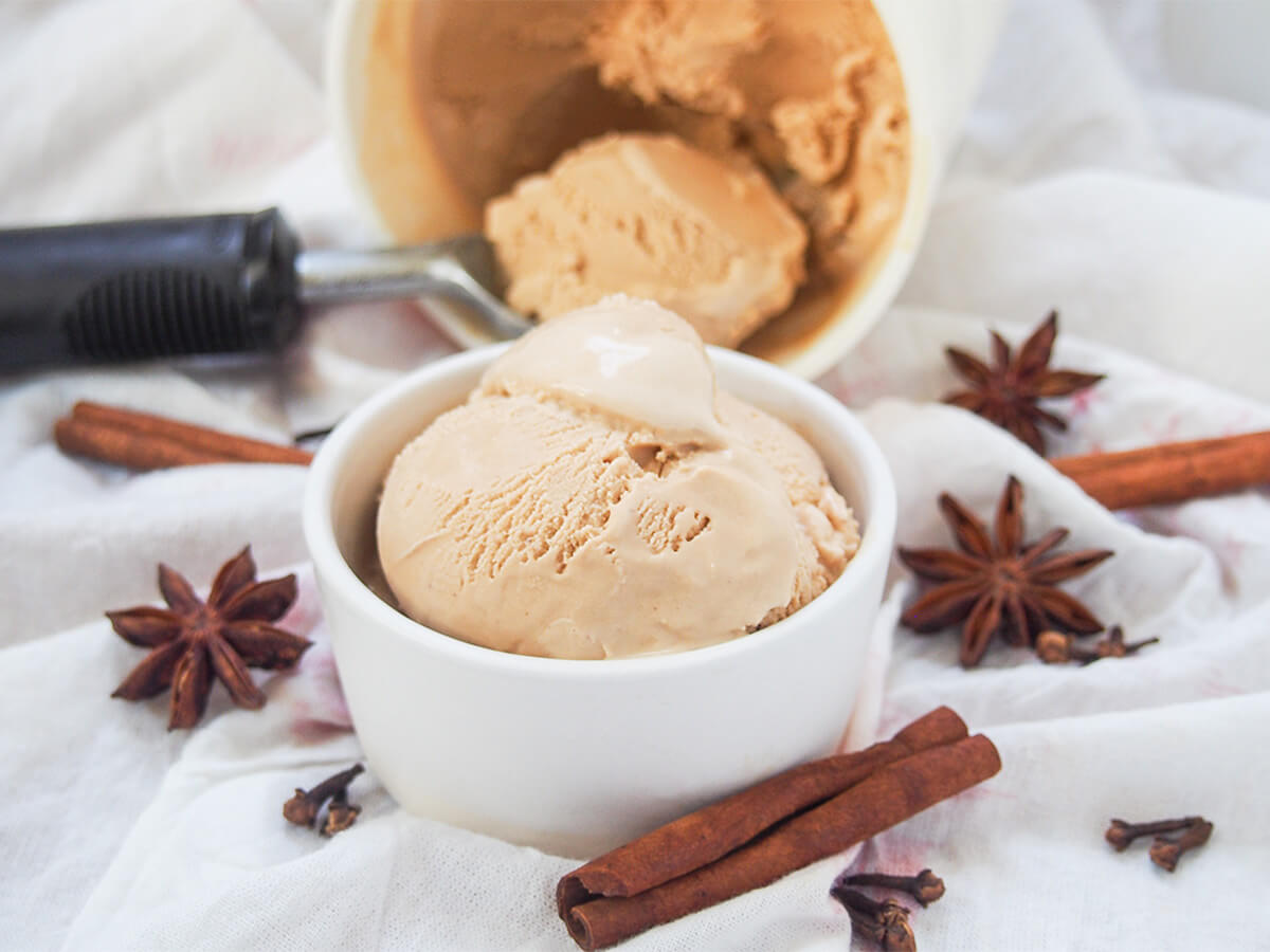 small bowl of chai ice cream with scoop in container behind and spices scattered either side.
