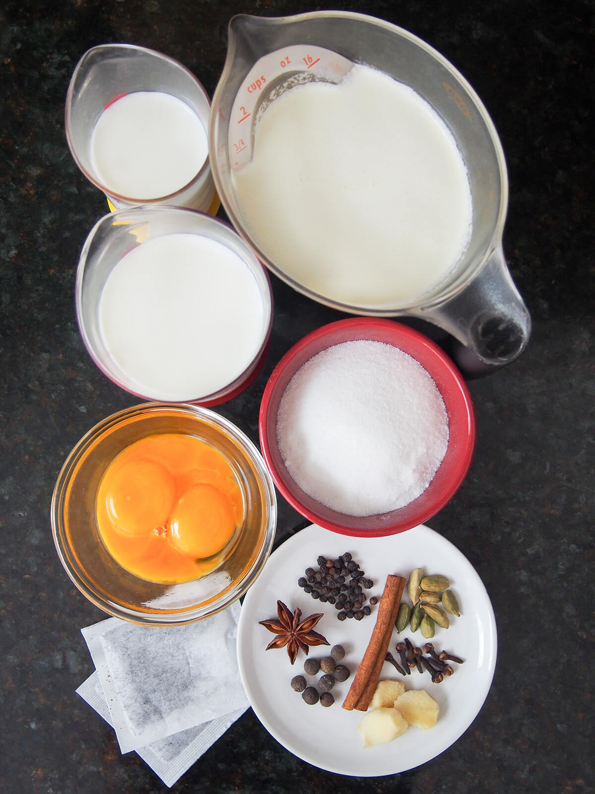 measures of milk, cream, bowls of sugar and egg yolks and plate with whole spices and tea bags below.