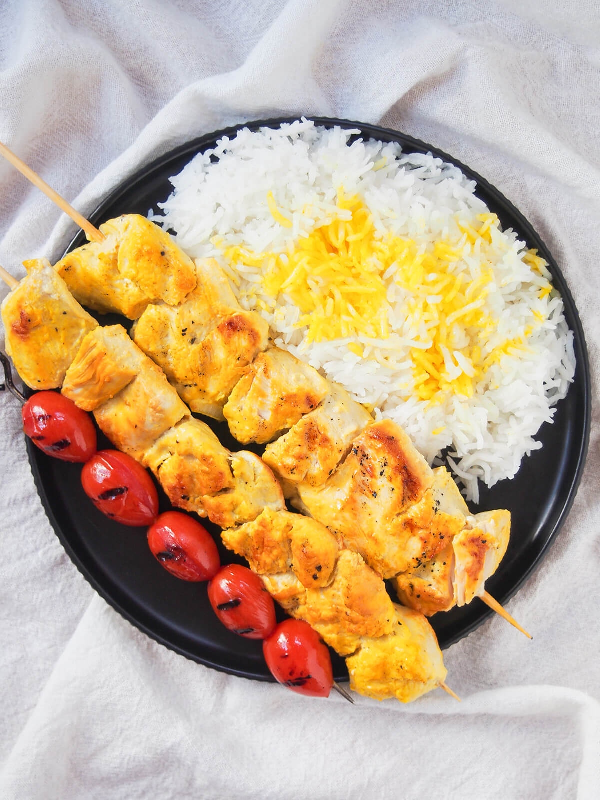 Joojeh kabab is an easy Persian skewer where chunks of chicken are marinated with saffron and yogurt. Easy, tender and delicious. plate with rice, two jujeh kabab and skewer of grilled tomatoes from overhead.