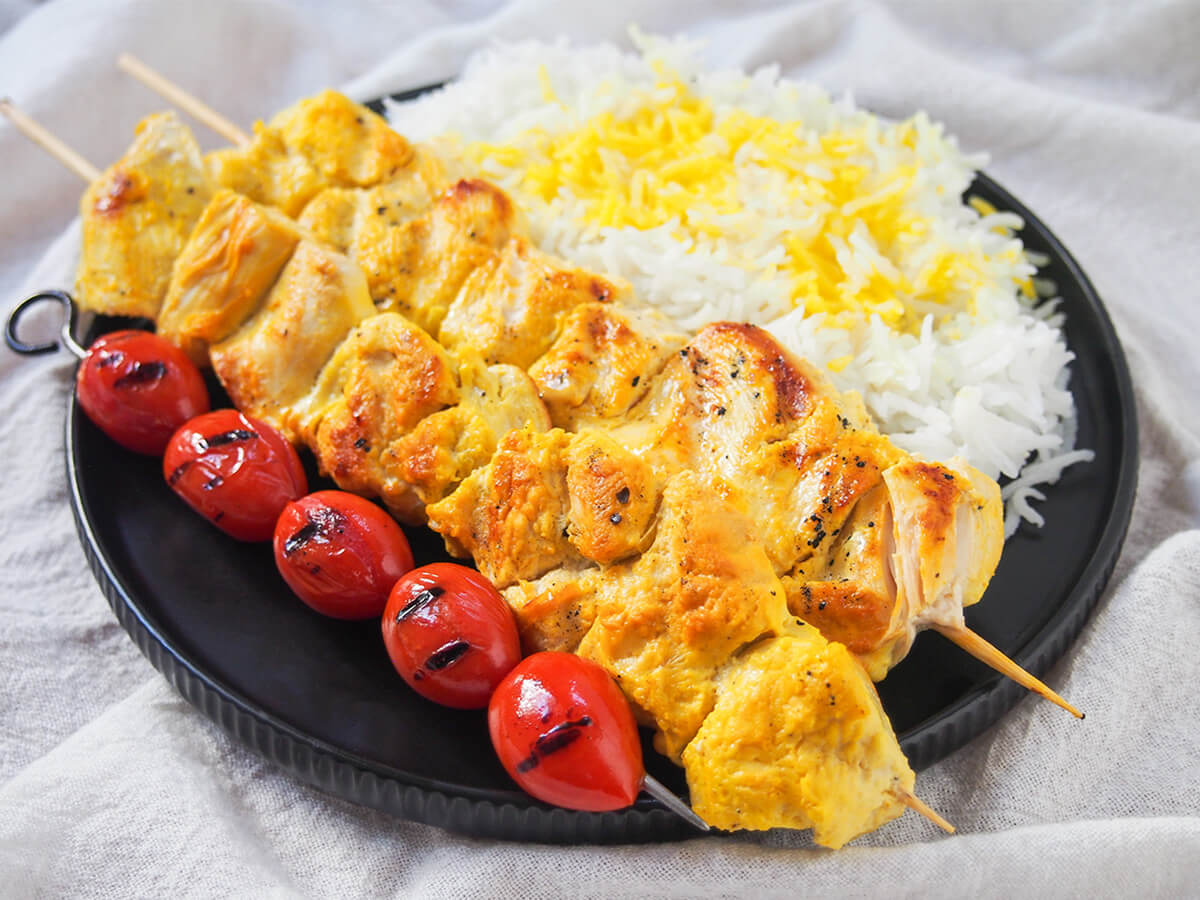 joojeh kabab skewers on plate with rice and grilled tomatoes jujeh kabab on plate with tomatoes in front, rice behind/