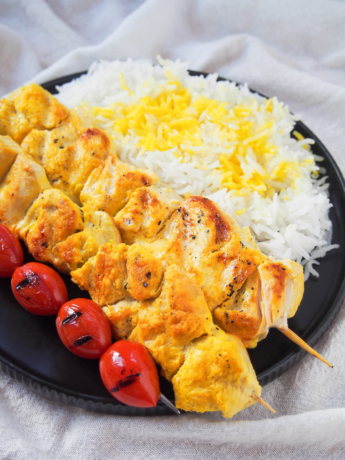 Joojeh kabab is an easy Persian skewer where chunks of chicken are marinated with saffron and yogurt. Easy, tender and delicious. partial side view of jujeh kabab, tomatoes and rice on plate.