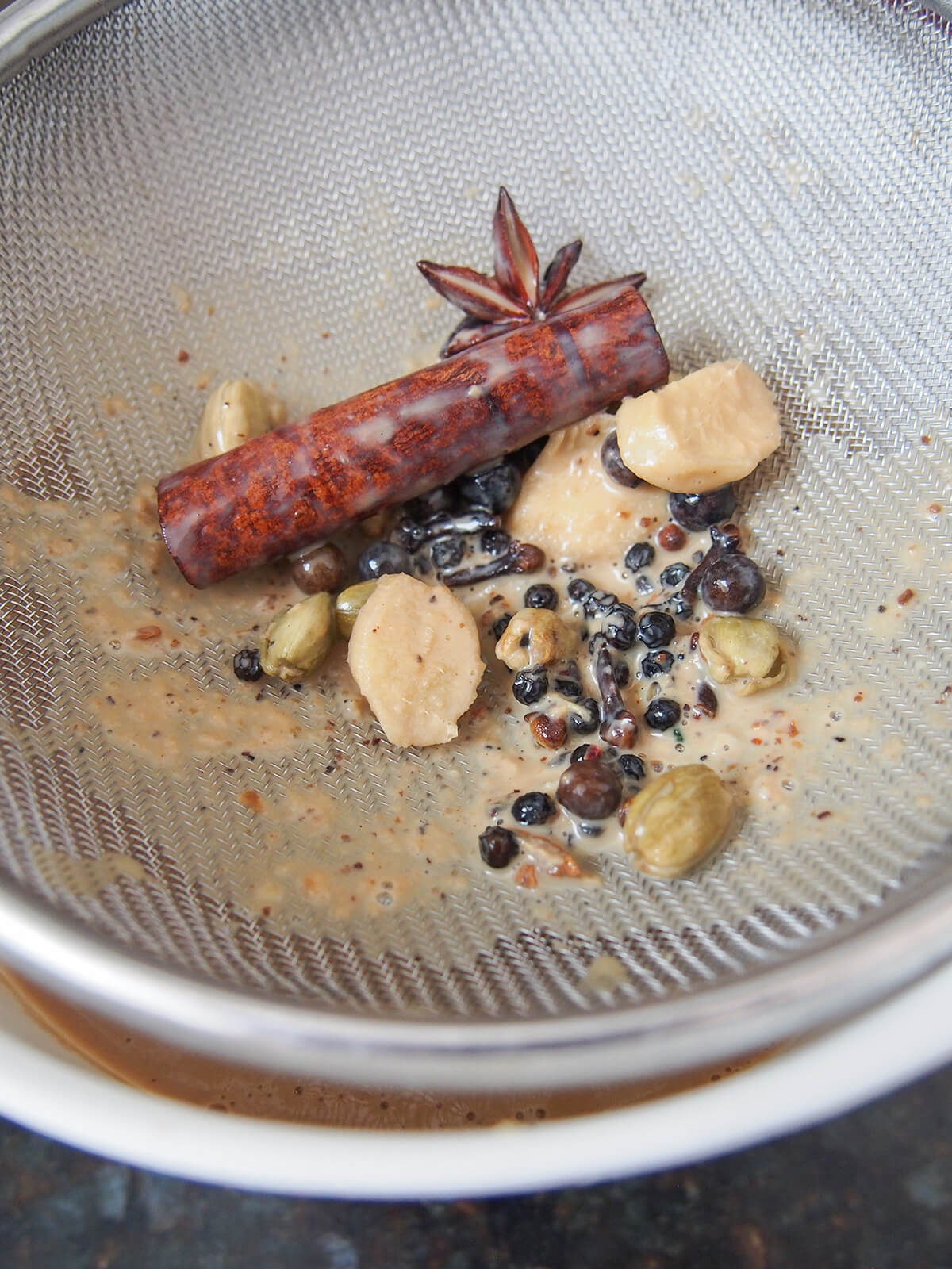 whole spices held back in strainer over bowl with brown liquid in just visible.