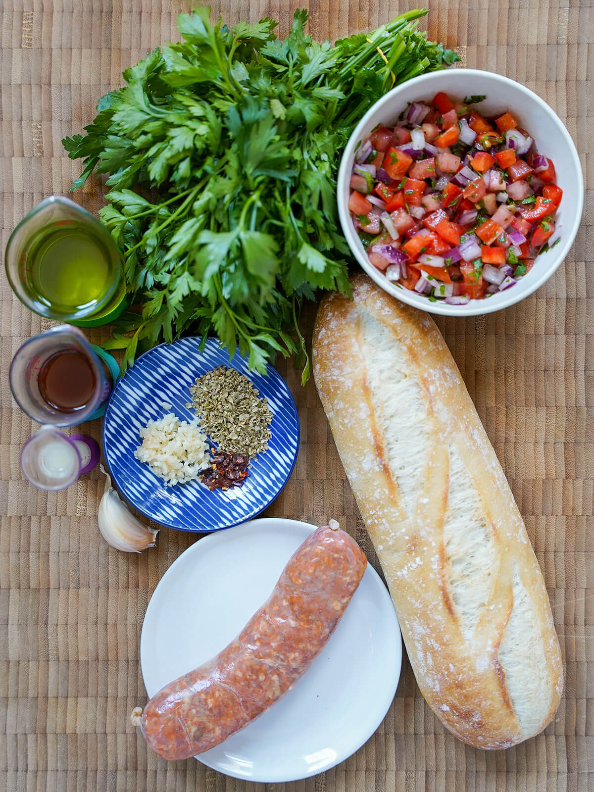 choripan Argentine chorizo sandwich ingredients chorizo sausage, demi baguette, bowl of salsa, bunch of parsley, oil, vinegar and small plate with oregano, garlic and chili.