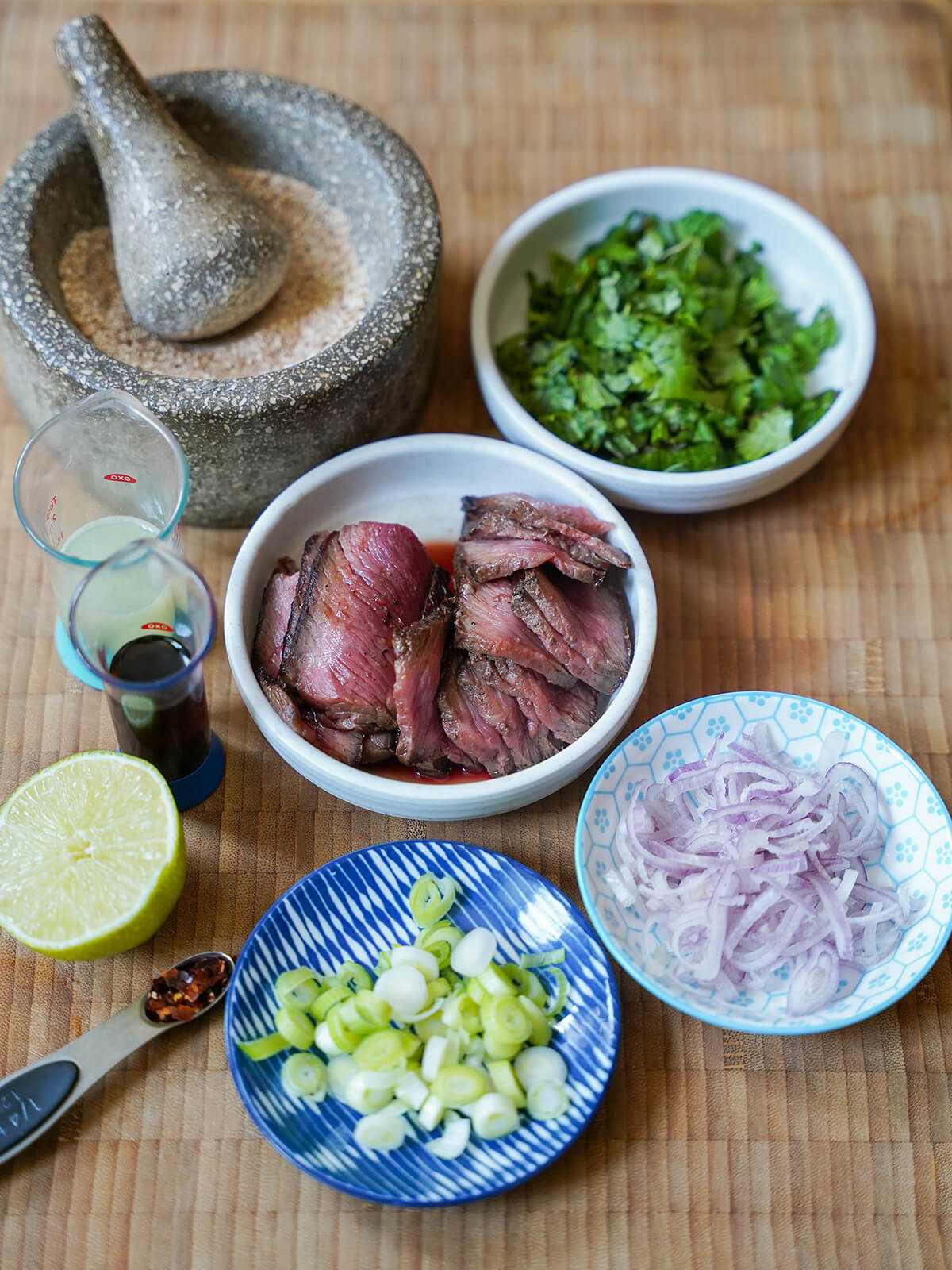 prepared ingredients for nam tok before combining plates of chopped herbs, sliced shallots and green onions, sliced beef with mortar of ground rice.