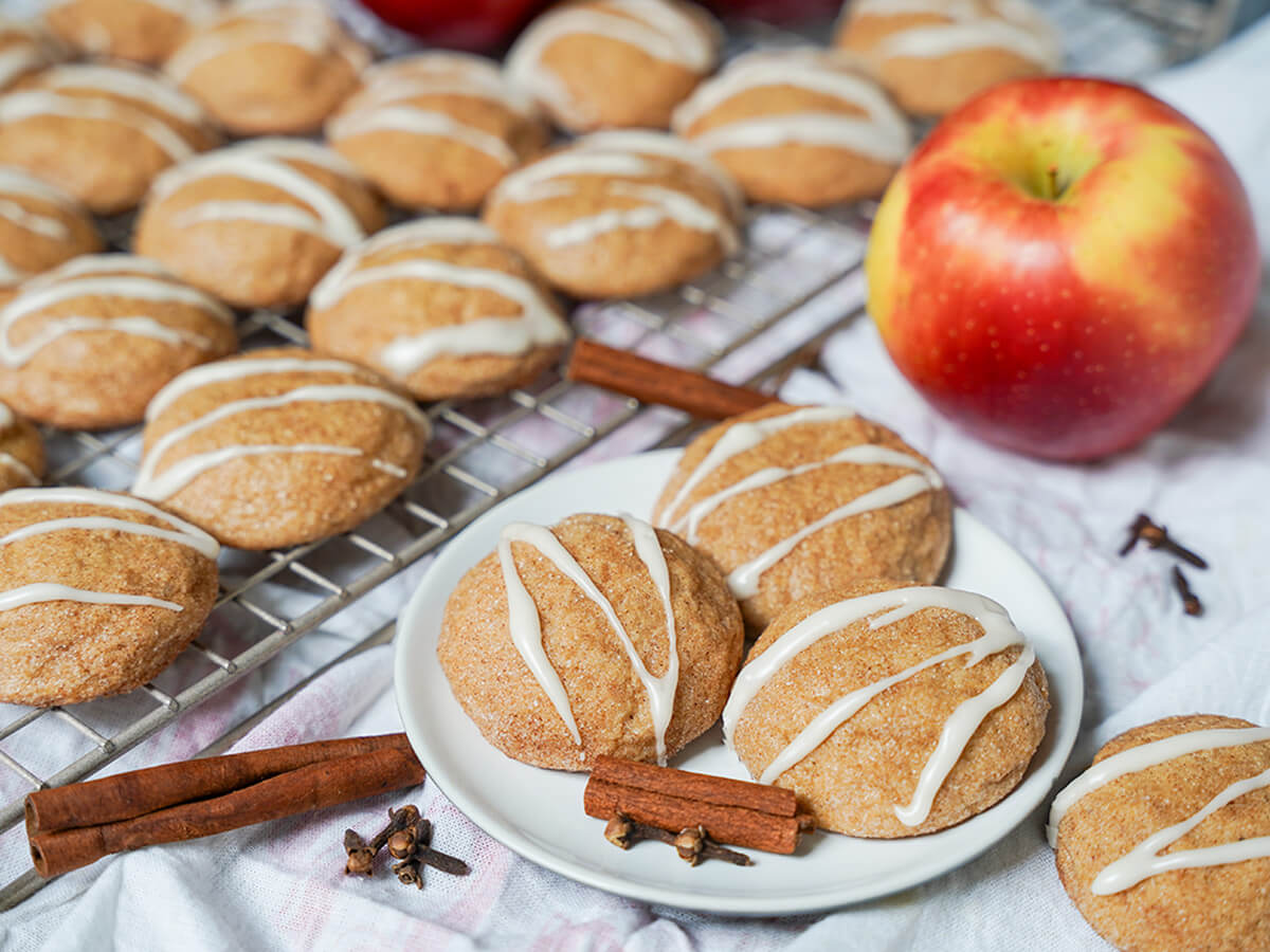 apple cider cookies plate of three apple cider cookies with more on cooling rack behind and spices and apple around.