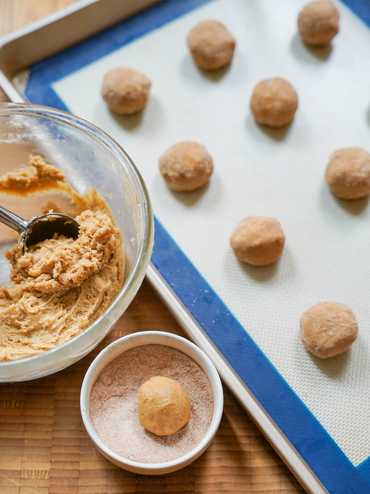 making apple cider cookies bowls of dough with scoop in it, a ball of dough in dish with cinnamon sugar and finished balls on lined baking sheet.