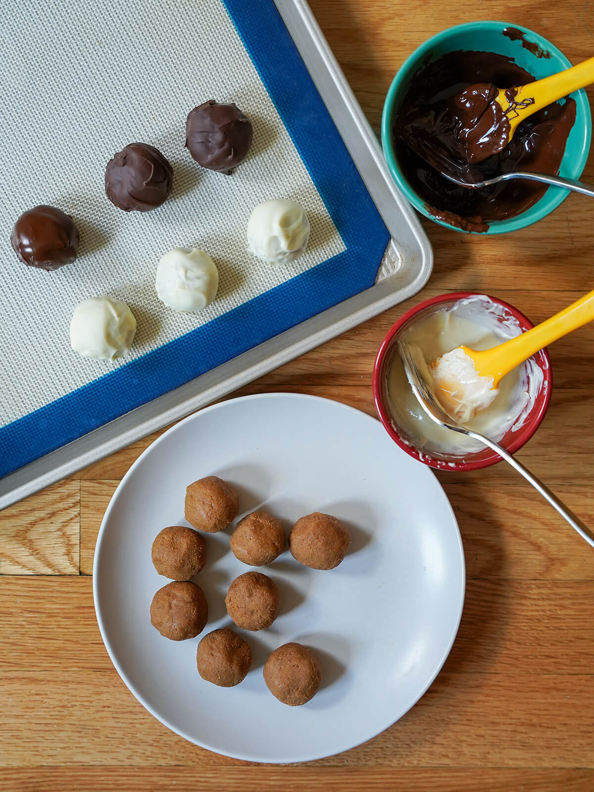 dipping balls of filling in chocolate are letting firm up for biscoff truffles balls of truffle filling on plate with bowls of melted chocolate and some dipped truffles on lined baking sheet.