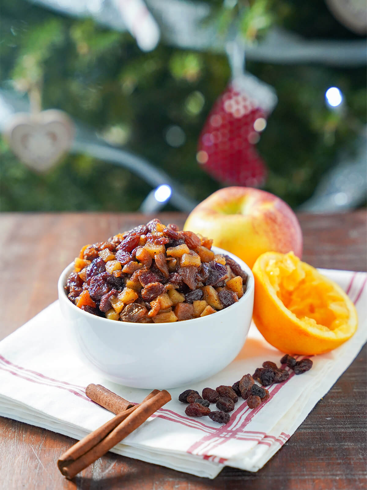 bowl of sugar free mincemeat with apple, orange, cinnamon stick and raisins around bowl and blurred tree in background.