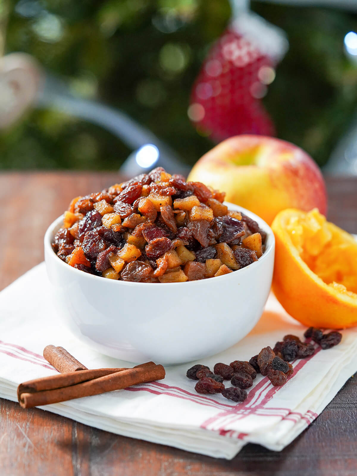 bowl of sugar free mincemeat with apple and squeezed orange behind and cinnamon stick and raisins in front of bowl.