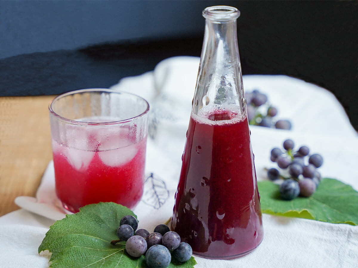 bottle of grape syrup with glass of grape soda to side with small bunches of grapes on leaves either side.