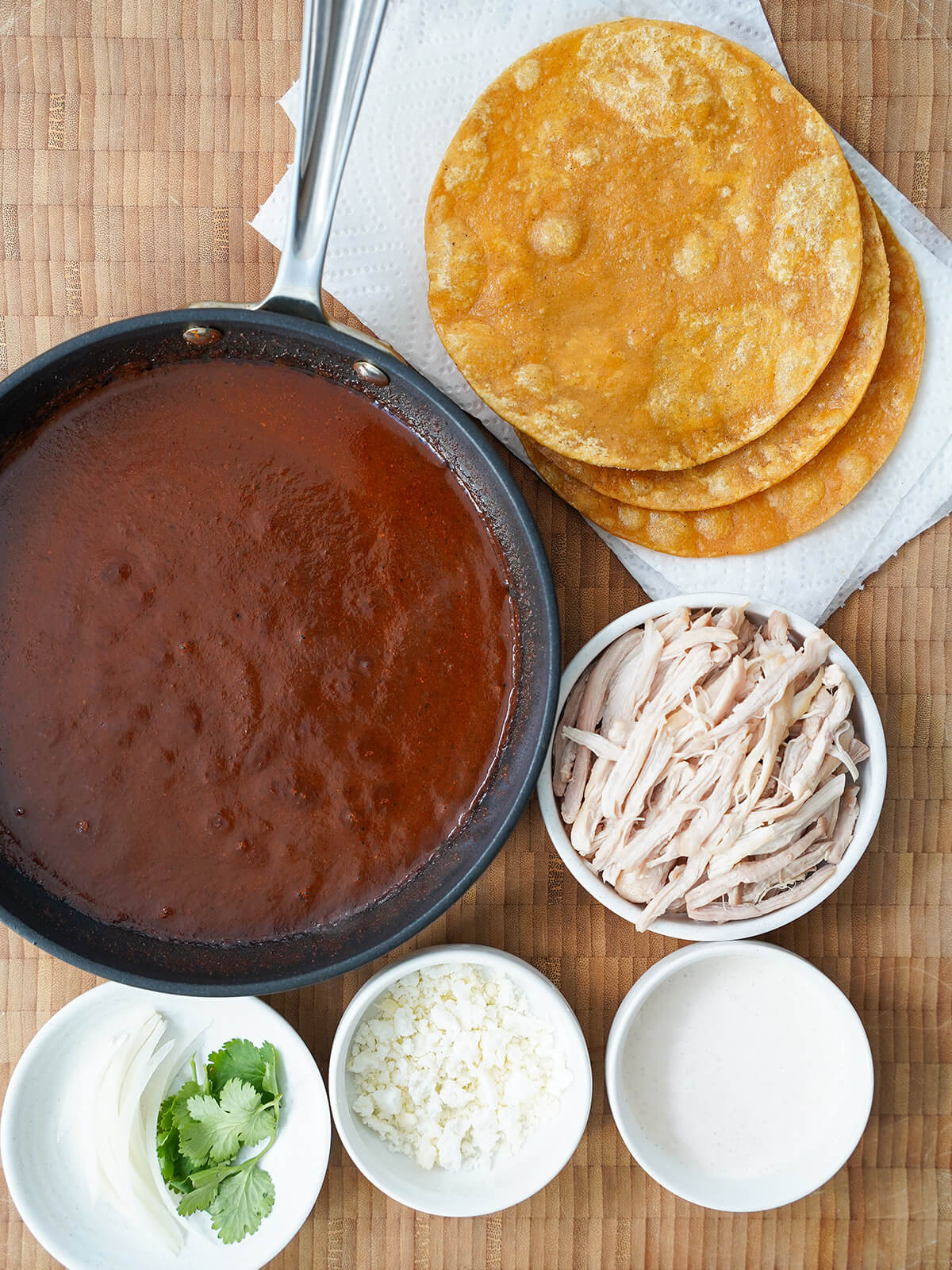 skillet of mole sauce, fried corn tortillas, dish of shredded chicken, dishes with crema, cheese and onion slices.