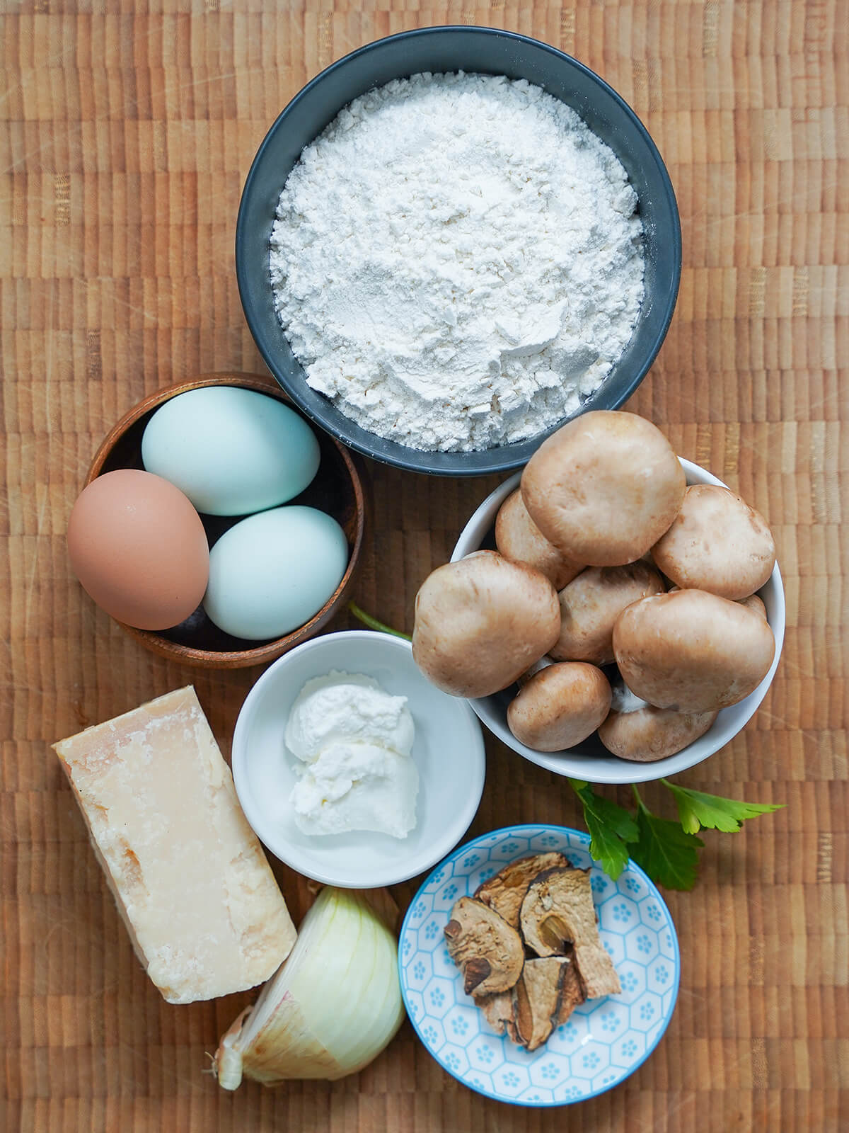 ingredients for mushroom ravioli bowls of flour, mushrooms, eggs, ricotta, dried porcini with onion and parmesan to side.