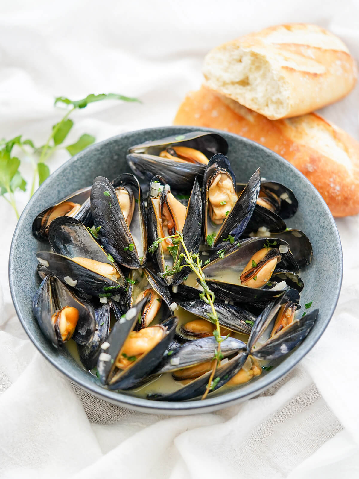 side view of mussels in cream with split baguette and parsley behind bowl.