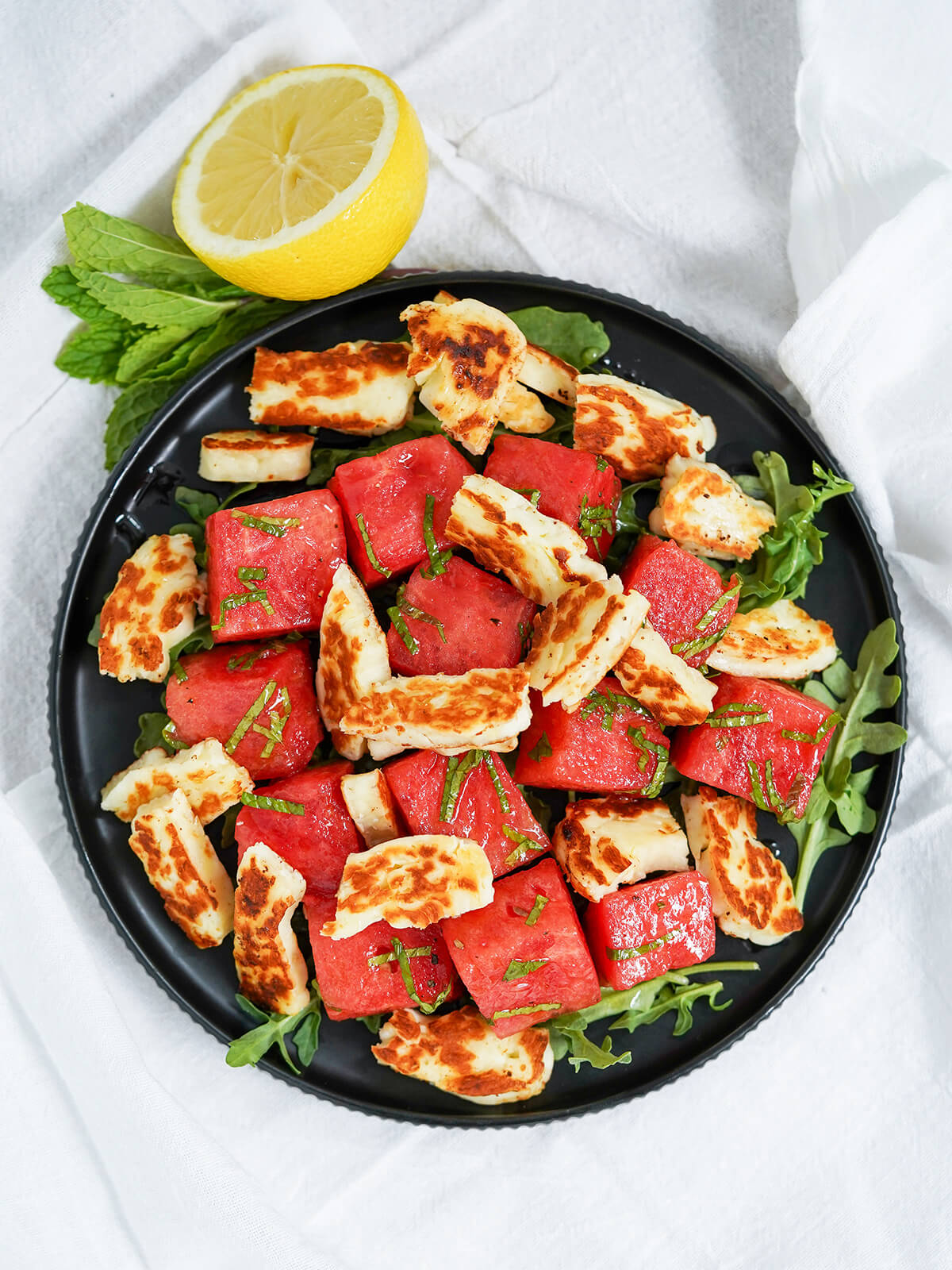 overhead view of plate of halloumi and watermelon salad with part lemon and sprig mint above plate.